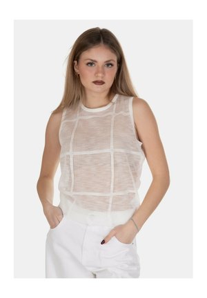 Giovane donna con capelli lunghi indossa una canottiera bianca in rete con motivo a griglia e pantaloni bianchi, in piedi con le mani nelle tasche.