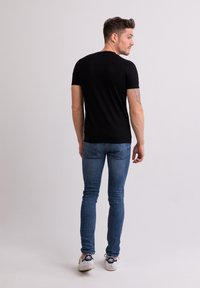 Černé tričko s krátkým rukávem v kombinaci s modrými džíny. Model stojí zády, ukazuje přiléhavou siluetu a neformální obuv.