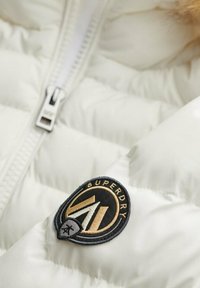 Hvid polstret jakke med en sølvfarvet lynlås og et sort rundt logo med guld accenter, der viser teksten "SUPERDRY."