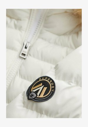 Chaqueta acolchada blanca con cremallera plateada y un logo circular negro con detalles dorados que muestra el texto "SUPERDRY."