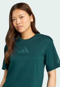 Tmavo zelené tielko s krátkym rukávom, okrúhlym výstrihom a jemne štruktúrovaným logom na hrudi. Hladký povrch látky.