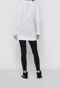 Maglione lungo bianco a maglia con un design ampio e aperto, dotato di maniche lunghe e orlo arrotondato. Abbinato a leggings neri e sneakers nere.