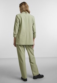 Blazer vert clair et pantalon assorti, présentant une coupe décontractée. Le blazer comporte une seule couture au dos ; le pantalon a des jambes larges. Chaussures noires.