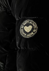 Sort dunjakke stof med en glat tekstur, der har et rundt, broderet mærke med et hjerte og tekst i guld og sort på ærmet.