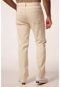 Beige Baumwollhosen mit einer körpernahen Silhouette, die über Gesäßtaschen, sichtbare Nähte und ein leicht zulaufendes Bein verfügen, kombiniert mit weißen Schuhen.