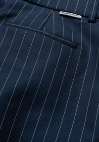 Gros plan sur un tissu bleu marine à fines rayures avec une poche passepoilée horizontale et une étiquette blanche portant la mention "ARMEDANGELS" près de la taille.