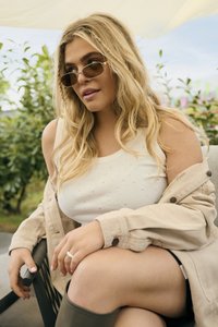 Joven mujer con cabello largo y rubio, usando gafas de sol, una blusa blanca sin mangas, chaqueta beige y botas grises, sentada al aire libre con vegetación detrás.