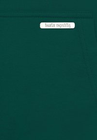 Green fabric with a smooth texture, featuring a silver metal tag embossed with "bazix republiq." Simple, uniform stitching detail.