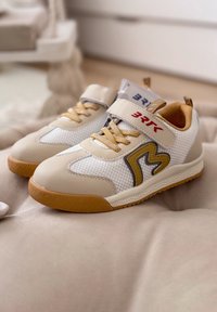 Paire de baskets pour enfants beiges et blanches avec une bride Velcro, semelle marron, et un "M" décoratif sur le côté, reposant sur une surface douce.