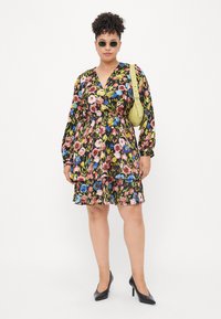 Floral dress with long sleeves, black base, pink, blue, and green patterns, cinched waist, tiered skirt, paired with black heels and green bag.