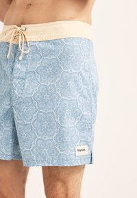 Bañadores de color azul claro con un patrón floral, con una cintura y cordón en color crema, y una etiqueta de logo en el dobladillo inferior.