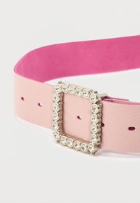 Ροζ δερμάτινη ζώνη με λεία επιφάνεια, τετράγωνη μεταλλική αγκράφα διακοσμημένη με μικρές, διαφανείς πέτρες και πολλαπλές τρύπες ρύθμισης.