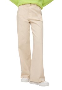 Beżowe, bawełniane spodnie dzwony z zapięciem na guzik, z dwoma przednimi kieszeniami i delikatną teksturą. Noszone z białymi sneakersami i zielonym swetrem.
