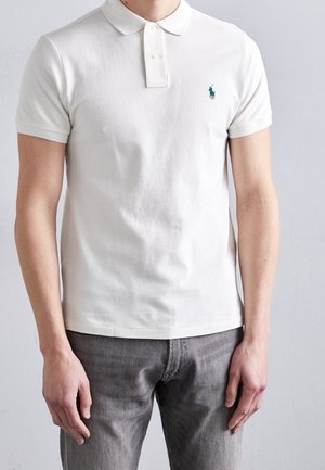 Homme portant un polo blanc à manches courtes avec un petit logo vert brodé sur la poitrine et un pantalon gris.