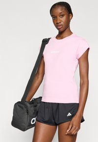 Ružové bavlnené tričko s vyhrnutými rukávmi a bielym logom "adidas", skombinované s čiernymi šortkami a čiernou športovou taškou s bielym logom.