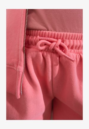 Lyserød fleece zip-jakke og matchende joggers med elastisk talje og snøre. Glat tekstur med en ensartet farve gennemgående.