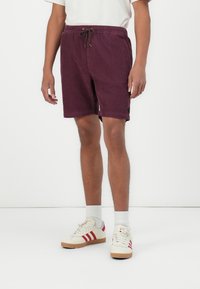 Bordeaux shorts met een elastische tailleband en trekkoord. Heeft zijzakken en een klein logo-detail. Draagt het met witte sneakers en sokken.