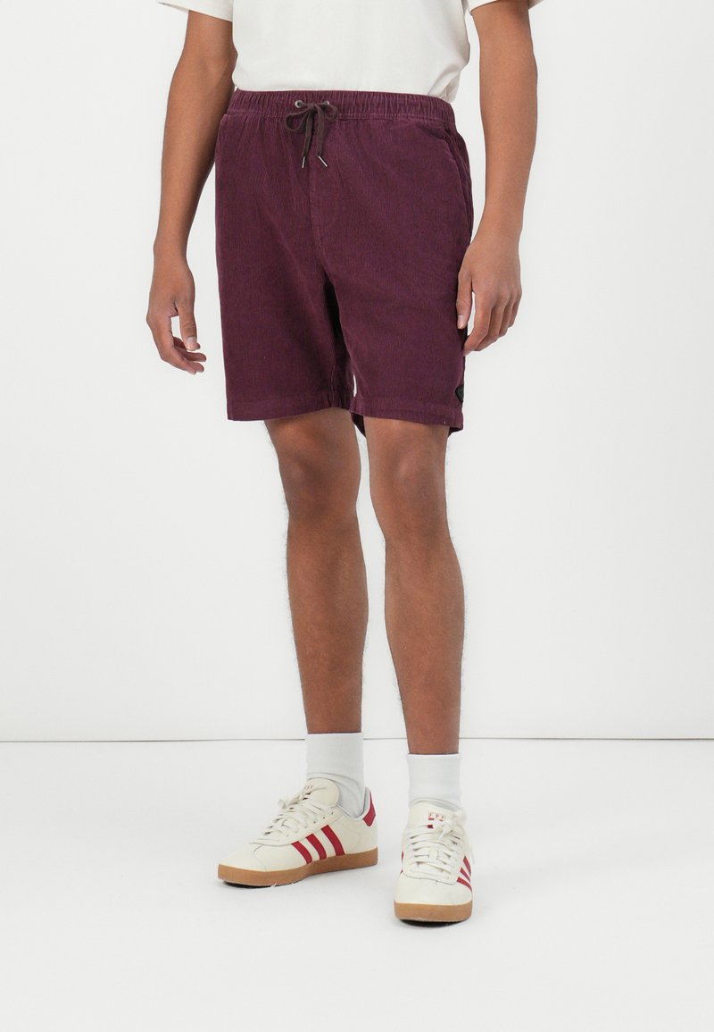 Bordeaux shorts met een elastische tailleband en trekkoord. Heeft zijzakken en een klein logo-detail. Draagt het met witte sneakers en sokken.