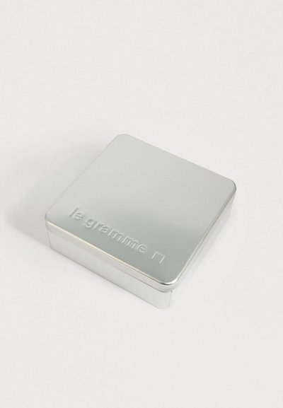 Caixa retangular de metal prata com cantos arredondados e o texto em relevo "le gramme" na tampa, colocada sobre uma superfície branca.