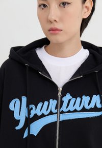 Sort zip-up hoodie i sort med lyseblå broderet tekst "Din tur" på brystet, med snoretræk i hætten og en hvid skjorte under.