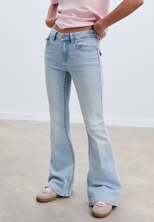 Helle blaue Schlagjeans mit mittelhoher Taille, ausgestattet mit Vordertaschen, Gürtelschlaufen und einem ausgewaschenen Finish. Kombiniert mit beigen Sneakers.