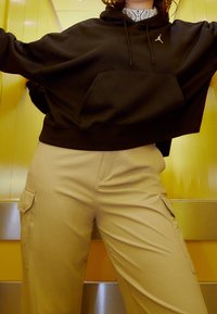 Sudadera negra recortada con bolsillo frontal y logo; pantalones cargo beige con bolsillos laterales, sobre un fondo amarillo.