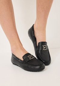 Svarta läderloafers med platt sula, med en dekorativ metallemblem på framsidan och silverfärgade detaljer.