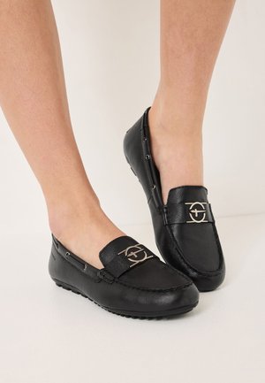 Schwarze Lederloafer mit flacher Sohle, ausgestattet mit einem dekorativen Metallsymbol auf der Vorderseite und silbernen Hardware-Akzenten.