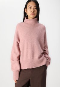 Rosa Turtleneck-Pullover aus weichem, flauschigem Material. Verfügt über einen gerippten Saum und Ärmelbündchen sowie eine lockere Passform. Kombiniert mit braunen Hosen.