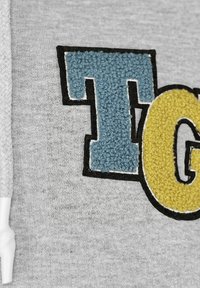 Grått tyg med en texturerad, broderad bokstav "T" i blått och "G" i gult, kantad av svart söm. Mjuk, stickad textur.