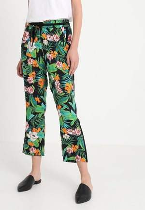 Mujer que lleva pantalones negros recortados con estampado floral tropical colorido y zapatos negros sin cordones, de pie sobre fondo blanco.