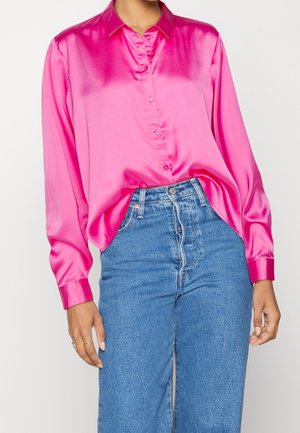 Woman wearing bright pink satin button-up shirt tucked into high-waisted blue denim jeans, standing against a plain background.