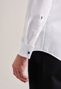 Mano maschile accanto alla manica di una camicia bianca con ricamo di rosa nera sul polsino e sulla patta della manica, abbinata a pantaloni neri.