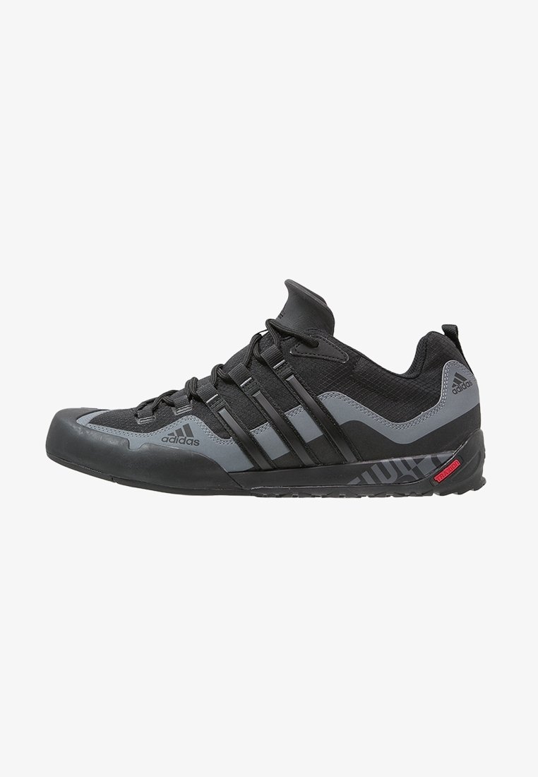 Chaussure de sport Adidas basse noire et grise avec lacets, semelle en caoutchouc texturée, logo latéral et embout renforcé.