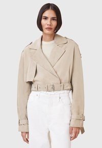 Beige Wildledercropped-Jacke mit breiten Revers, tailliertem Gürtel und knöpfbaren Manschetten. Trägt man über einem hellen Oberteil und kombiniert mit weißen Jeans.