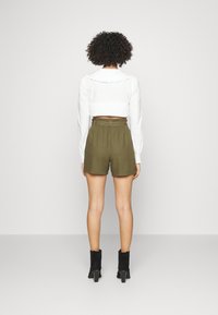 Camicia bianca a maniche corte con colletto arricciato, abbinata a pantaloni a pantaloncino verde oliva con un elastico in vita. La modella indossa stivali neri alla caviglia.