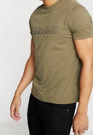 Mann trägt olivgrünes Kurzarm-T-Shirt mit „NAPAPIJRI“-Logo über der Brust und schwarze Hose, steht vor einfarbigem Hintergrund.