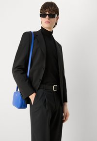 Zwarte op maat gemaakte colbert over een zwarte coltrui, gecombineerd met zwarte broek. Een kleine blauwe crossbodytas hangt aan een blauwe schouderband.