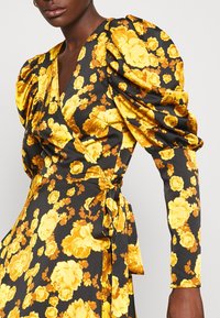 Femme portant une robe portefeuille noire à grosses manches bouffantes avec un motif floral jaune vif, nouée à la taille sur un fond blanc uni.