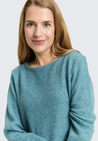 Vrouw met lang blond haar, gekleed in een blauwe gebreide trui, die vriendelijk glimlacht tegen een effen lichte achtergrond.