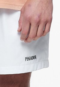 Bílé šortky vyrobené z lehkého materiálu s černým logem "PEGADOR" na lemu. Tkanina má hladkou texturu s ohrnutým lemem.