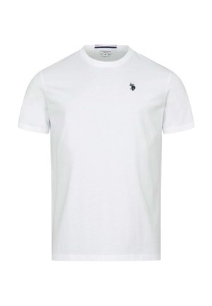 Mann lächelt und geht vorwärts, trägt ein weißes T-Shirt, schwarze Jeans und weiße Sneakers vor einem einfarbig hellen Hintergrund.