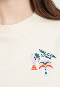 Βαμβακερό t-shirt σε κρεμ απόχρωση με γραφικό τύπωμα από χέρια και μια φοινικιά σε διάφορα χρώματα, με το κείμενο "VOLCOM STONE" από πάνω.