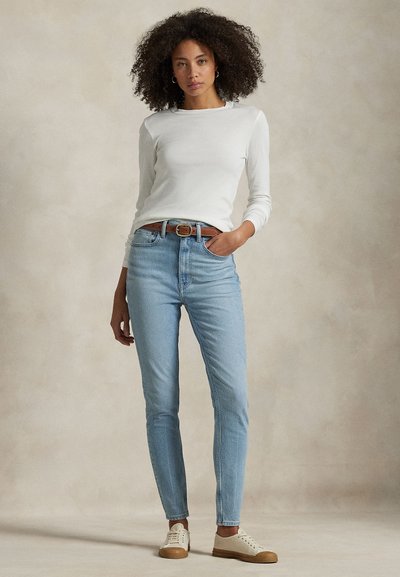 Vrouw met krullend haar die een wit shirt met lange mouwen draagt, lichtblauwe spijkerbroek met een bruine riem en witte sneakers, staand met één hand in de zak.