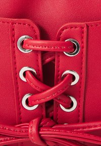 Corset en simili cuir rouge avec des œillets en métal argenté et des lacets. Couture détaillée et un accent de lacet noué en bas. Texture lisse.