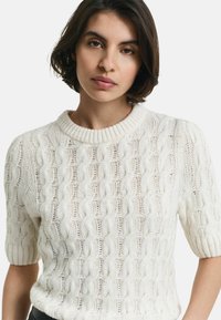 Gebreide trui in off-white met korte mouwen, voorzien van een gestructureerd kabelpatroon en een geribbelde ronde hals. Het materiaal lijkt zacht en ademend.