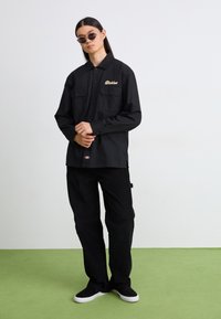 Camicia nera con bottoni, due tasche sul petto, testo ricamato in oro, abbinata a pantaloni neri e scarpe nere. Tessuto morbido, vestibilità comoda.