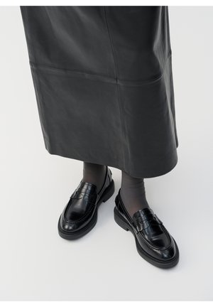 Czarne skórzane loafers noszone z ciemnymi przezroczystymi rajstopami i czarną skórzaną spódnicą midi na białym tle.