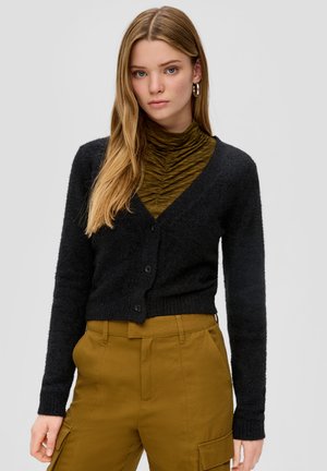 Cardigan noir et duveteux avec un décolleté en V profond, fermeture à boutons et manches longues, associé à un col roulé texturé vert olive et à un pantalon beige.