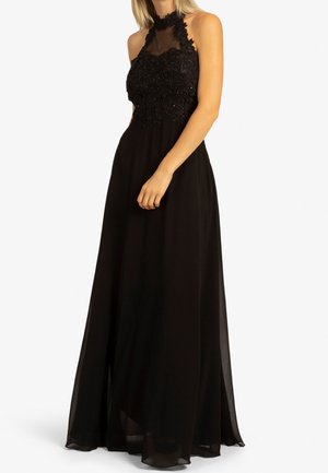 Femme portant une robe de soirée noire sans manches avec un corsage en dentelle et un col montant, debout devant un fond blanc uni.